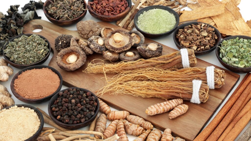 Adaptogens for Stress and Energy Balance 1 A wooden cutting board displays an assortment of dried herbs, spices, and medicinal roots. Prominent items include bundled ginseng roots, shiitake mushrooms, cinnamon sticks, turmeric rhizomes, and small bowls containing powdered spices (red, green, brown), whole peppercorns, and dried leaves. Scattered around are ginger slices, licorice root, and other botanicals, all arranged against a clean white background.