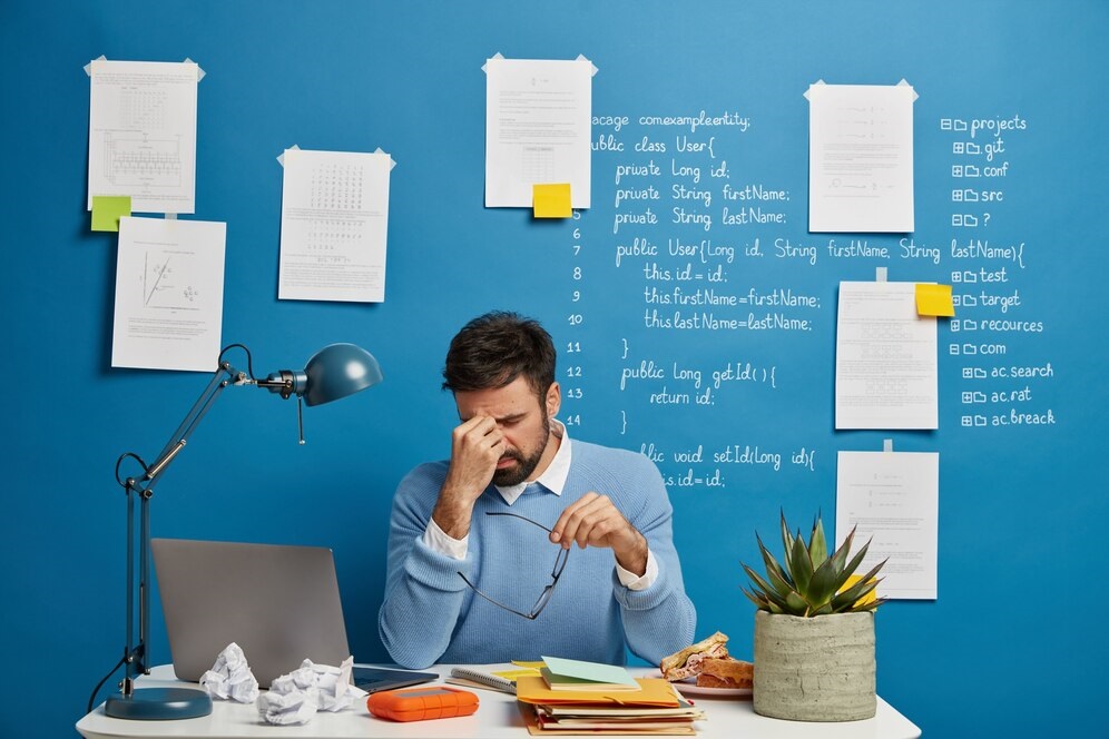 A stressed male programmer in a light blue sweater sits at a cluttered desk with a laptop, crumpled papers, a half-eaten sandwich, and a spiky green plant. Behind him, a blue wall is covered in handwritten Java code (e.g., 'public class User'), project file extensions (.git, .conf), and pinned notes, reflecting intense coding work.