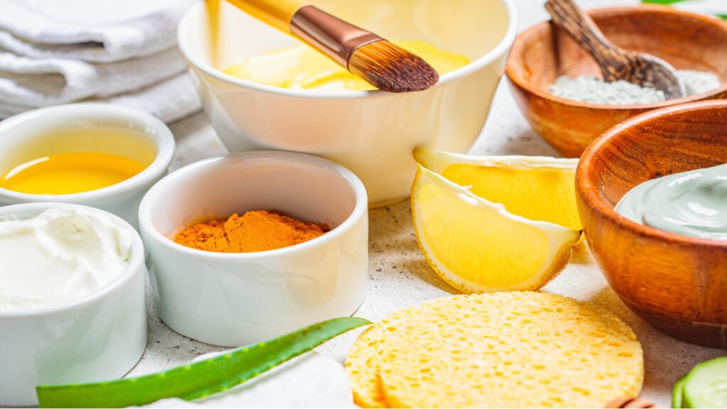 Natural Skincare Protocols for Eczema: Turmeric powder, lemon slices, aloe vera, yogurt, honey, green clay, and sponge in white and wooden bowls on countertop.