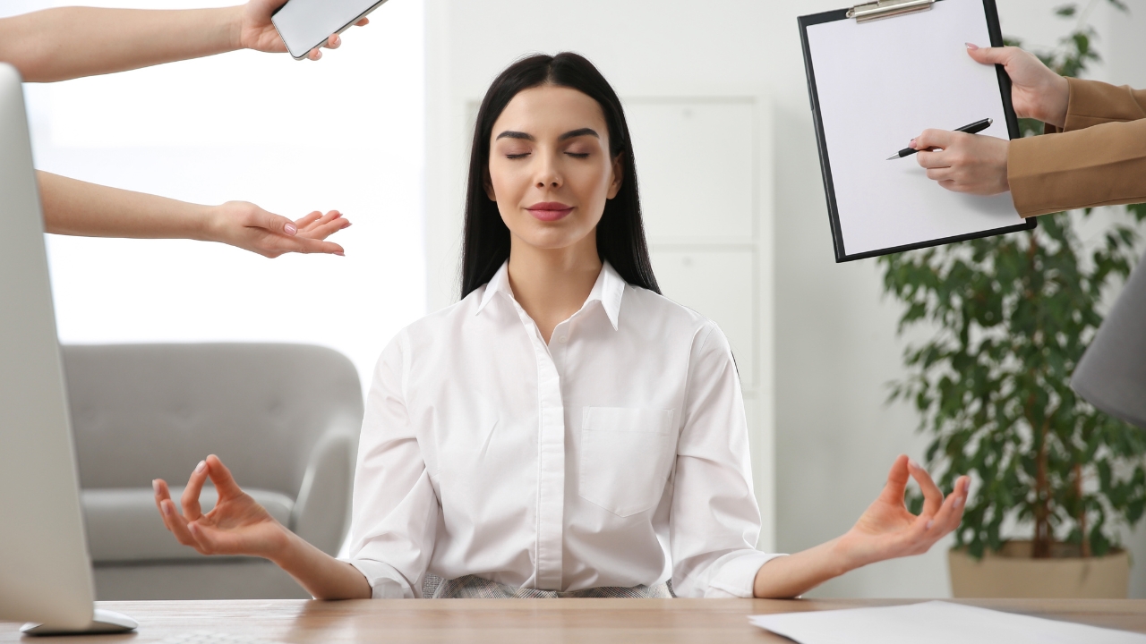 A calm woman meditates at her desk in an office, eyes closed and hands resting in her lap, surrounded by multiple hands reaching towards her—one holding a smartphone and another a clipboard—symbolizing workplace distractions.